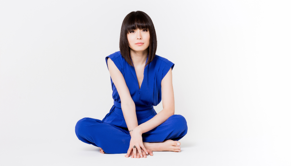 Alice Sara Ott stares into the camera. She's wearing a bright blue jumpsuit and sitting cross-legged on a neutral white background