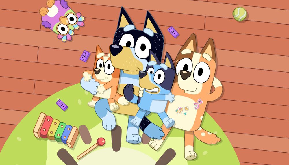 A cartoon dog family, two of which are blue and the other two are brown, lie on a green rug against a wooden floor gazing up at the sky. There is a pink owl toy, a ball and a xylophone on the rug with them.