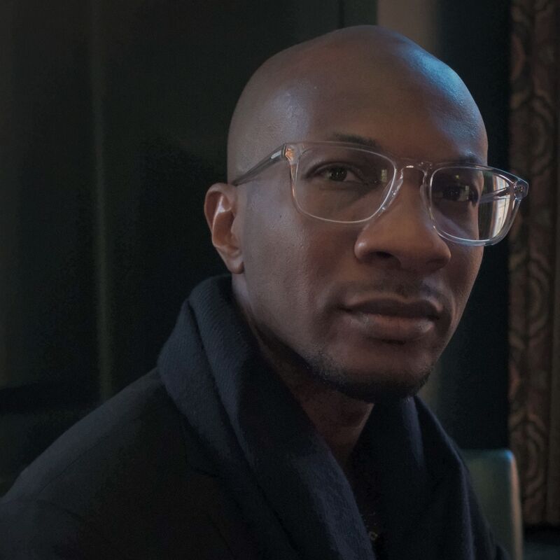 Photograph of Teju Cole wearing a dark jacket and glasses with clear frames.