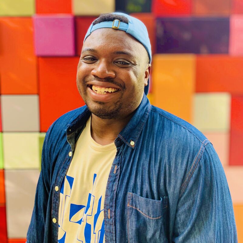 Bryan Washington wears a denim shirt with yellow tshirt underneath and a blue baseball cap. He is photographed against a red, pink and yellow patchwork background.