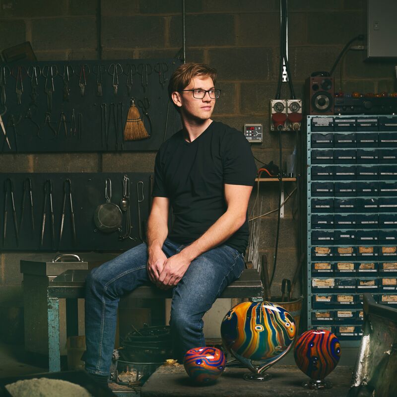 Will is pictured in a glass-blowing studio and wears dark blue jeans and a black t-shirt. He has light brown hair and black-framed square glasses.