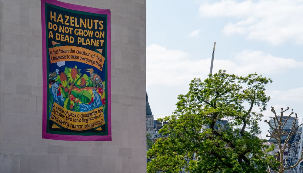 Ed Hall's artwork Hazelnuts do not grow on a dead planet, a brightly coloured banner work, in situ on the side of the Southbank Centre's Royal Festival Hall with trees visible in the background