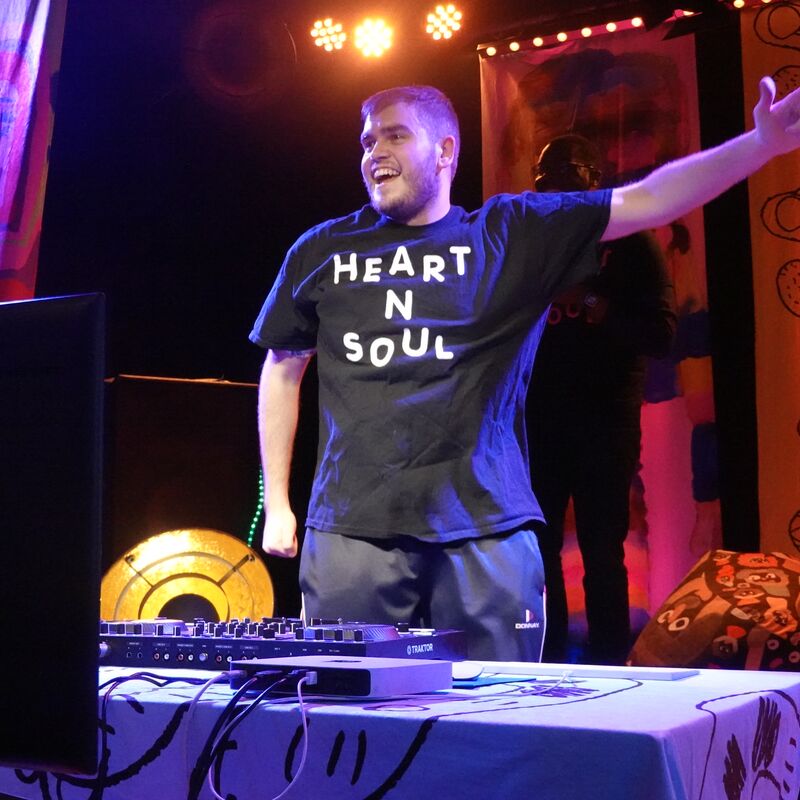 DJ with a black t shirt on stands with his hands up in the air in front of DJ decks.