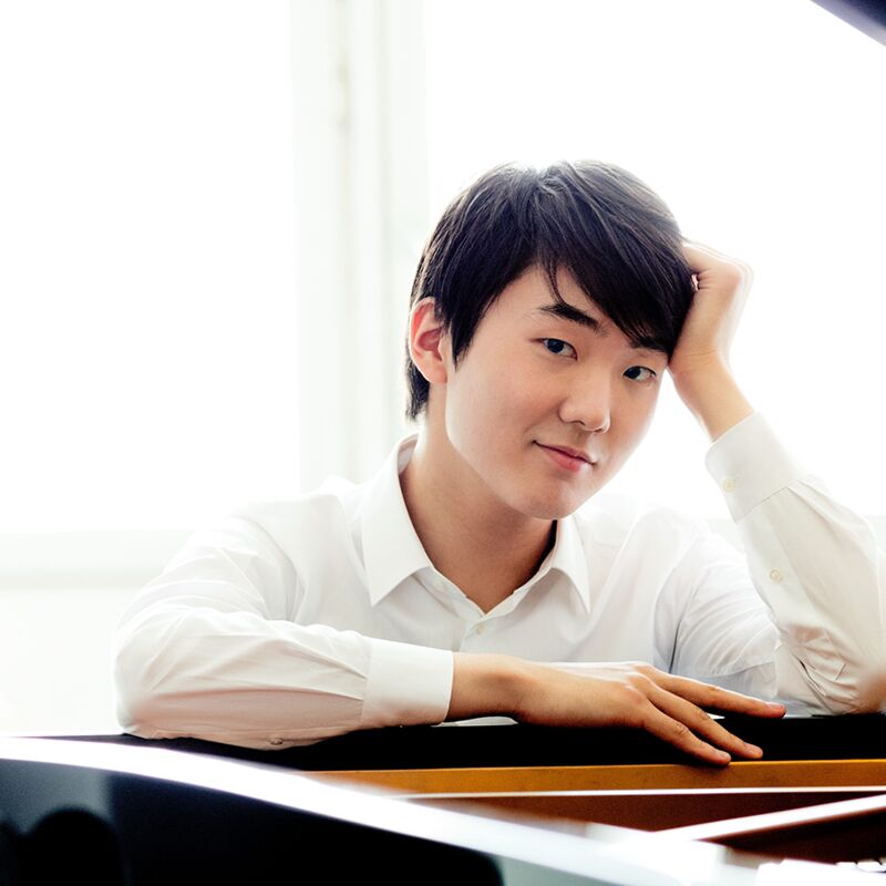 Seong-Jin Cho seated at a grand piano, smiling, in a white shirt