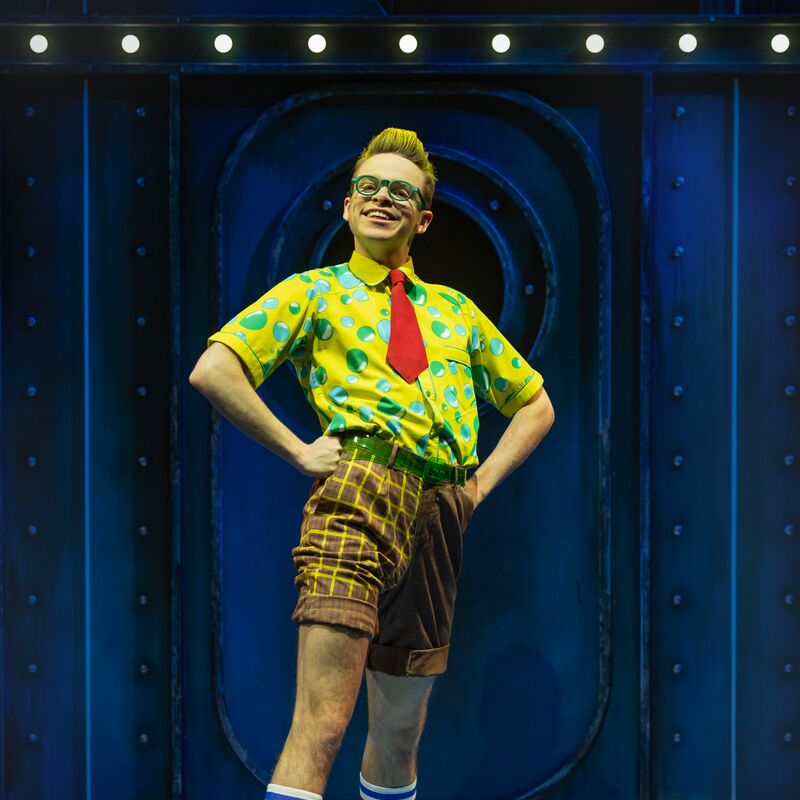 Lewis Cornay is shown playing SpongeBob SquarePants in The SpongeBob Musical. SpongeBob is pictured standing in front of a blue door. He has bright yellow hair and green-rimmed glasses, and is wearing a yellow shirt with a pattern of blue bubbles, a bright red tie and brown shorts. He is smiling and has his hands on his hips.