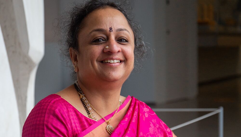 Supriya Nagarajan wearing a pink sari sitting down with a microphone and smiling.