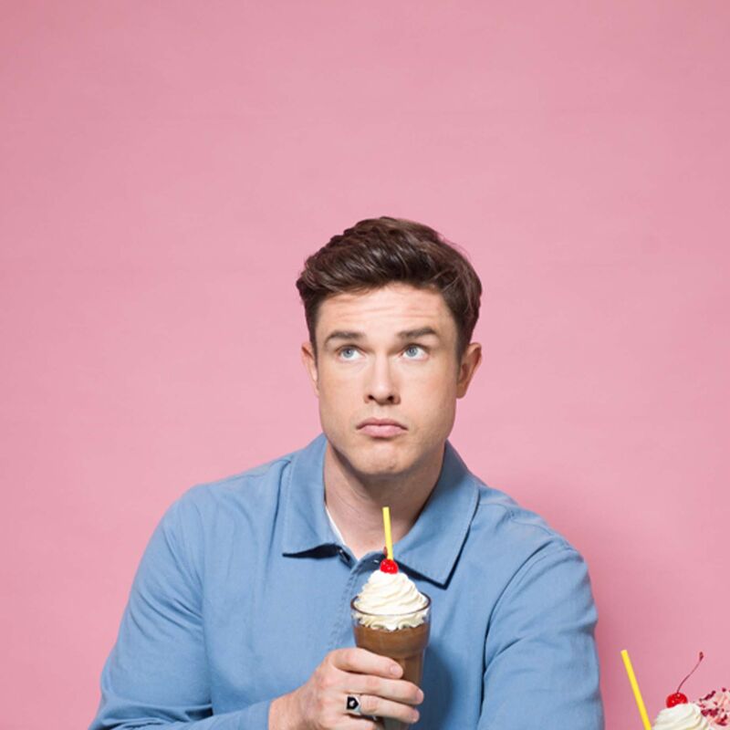 Comedian Ed Gamble sits infront of a full table of food holding an ice cream sundae and looking guilty.