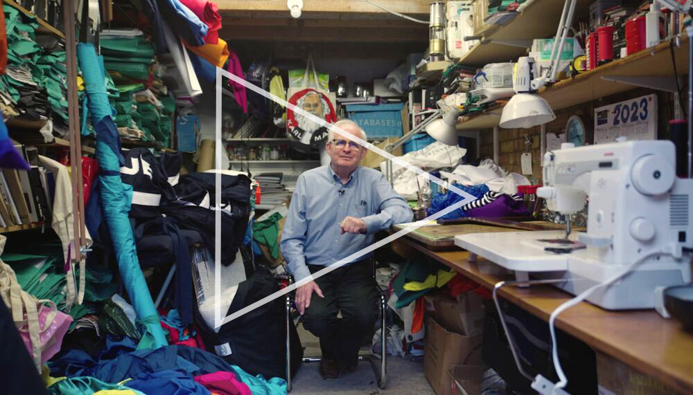 Banner-maker Ed Hall, an older White man with white hair, sits in his studio surrounded by cloth, banners and banner-making equipment