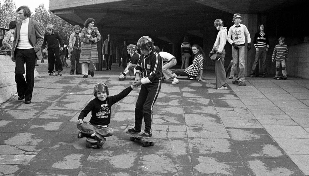 Young skateboarders in action at the undercroft skate space in 1976; in the foreground one skateboarder pulls along a nother one who is sitting on his board.