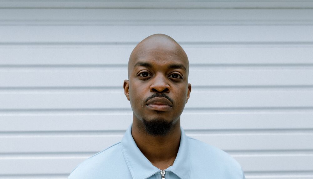 George the Poet wearing a blue shirt standing in front of a white corrugated background
