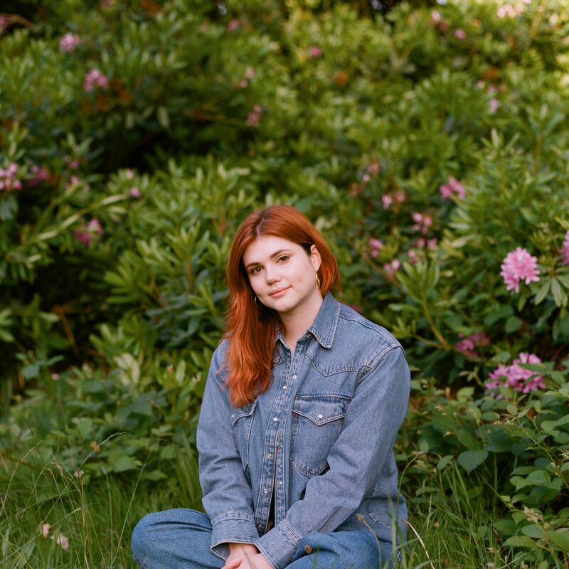 Eliza Clark is dressed in blue denim sitting in grass with flowers behind her