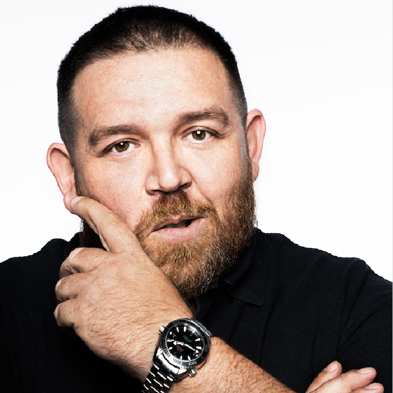 Nick Frost looks directly into camera. He has his arms folded and one hand up to his face, stroking his beard. He wears a black tshirt and has short dark brown hair.