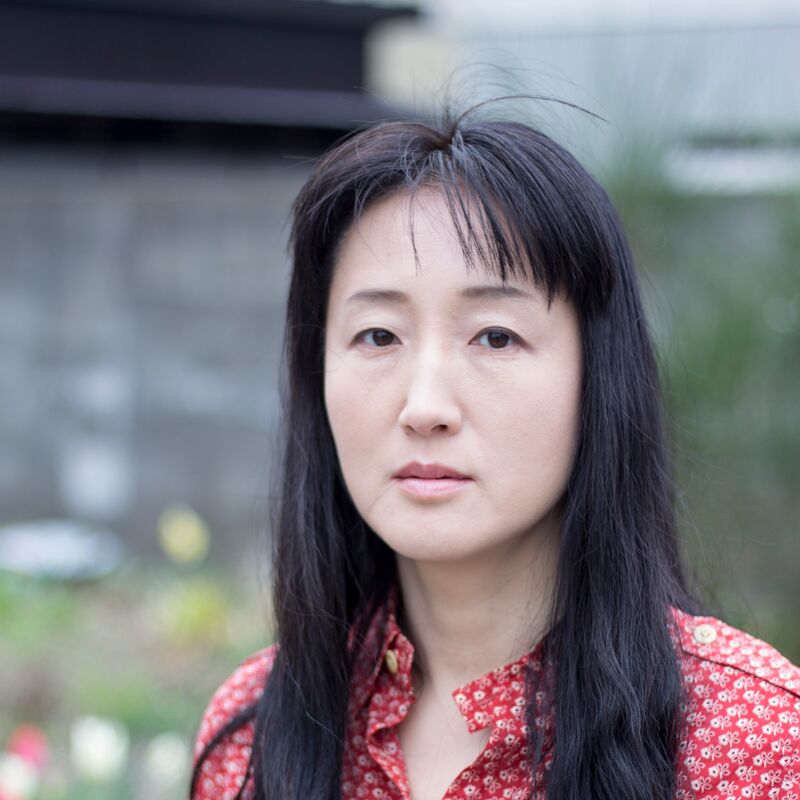 Headshot of Yu Miri in a garden, wearing a white and red blouse