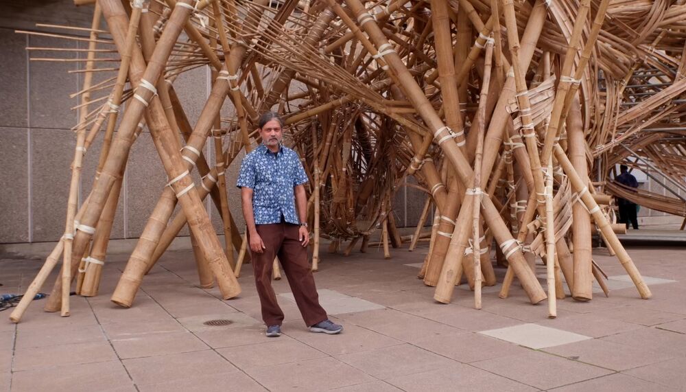 Asim Waqif, an Indian man with grey hair wearing a blue patterned shirt, stands in front of his Bagri Commissioned bamboo sculpture, वेणु [Venu], outside Hayward Gallery, 2023