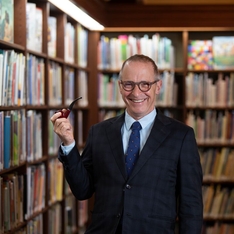 David Sedaris standing in a library smiling and holding a tobacco pipe
