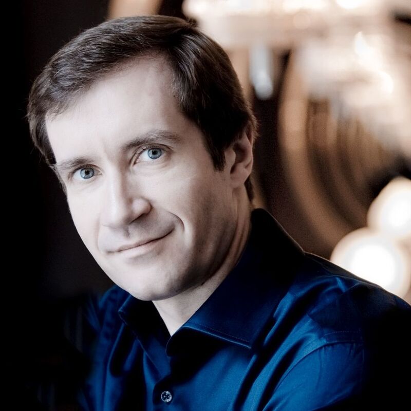 Head and shoulders portrait of pianist Nikolai Lugansky,. He looks directly into the camera with a soft smile, wearing a navy shirt