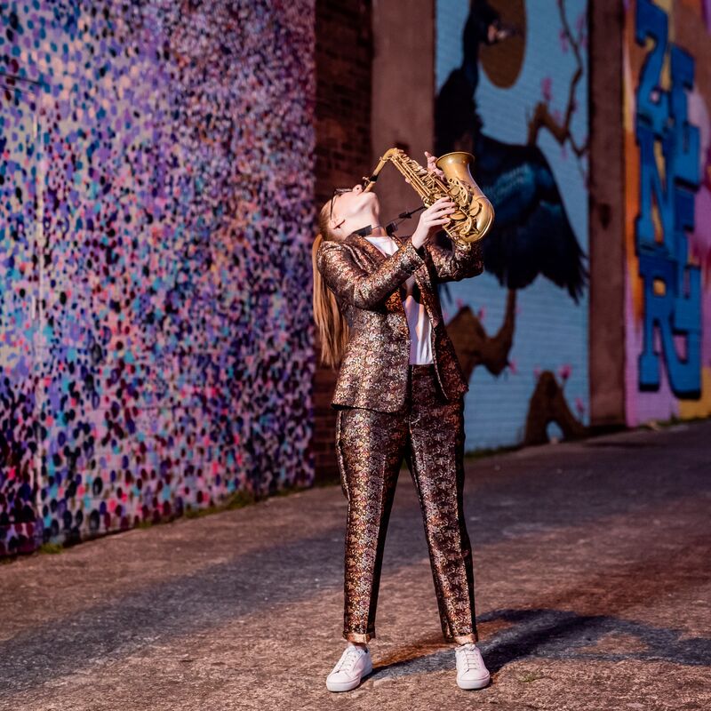 Full length portrait of Jess Gillam playing sax. She wears a bronze brocade suit and stands in a graffitied tunnel