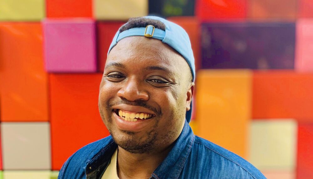 Bryan Washington wears a denim shirt with yellow tshirt underneath and a blue baseball cap. He is photographed against a red, pink and yellow patchwork background.