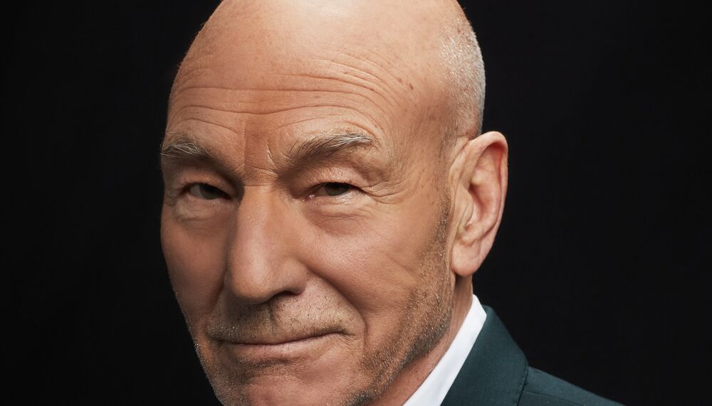 Patrick Stewart wears a sharp, dark suit over a white shirt and is pictured smiling to camera at a three quarter angle. He is bald with a faint stubble and dark eyes.
