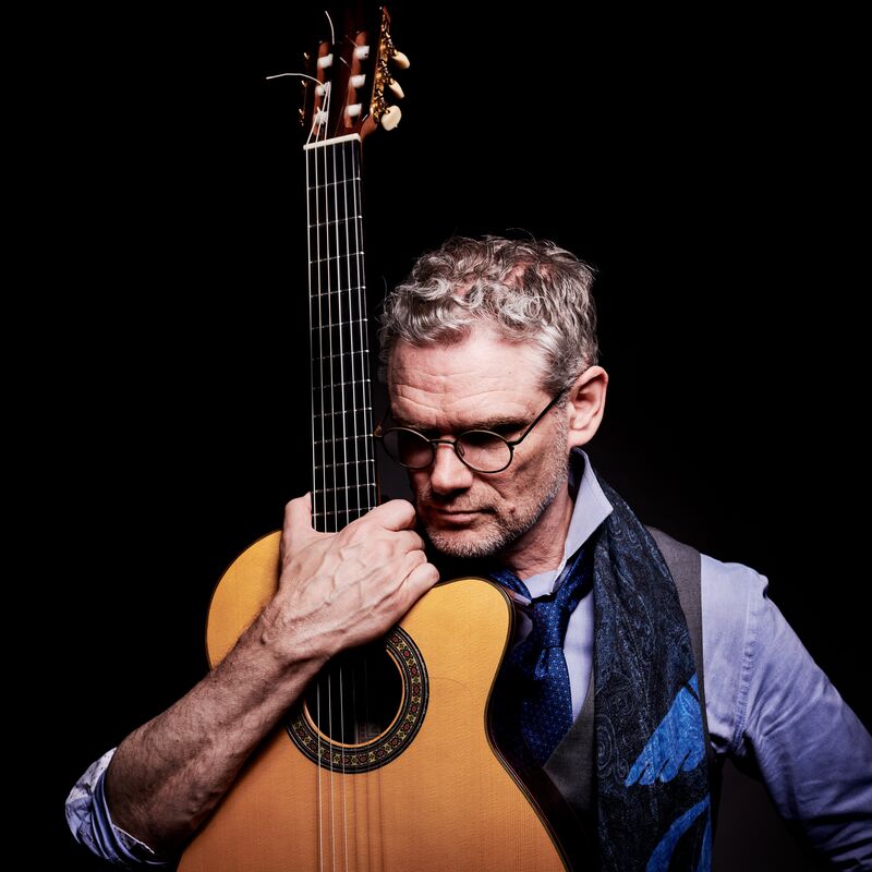 A man, Jesse Cook, holding a guitar looking away from the camera