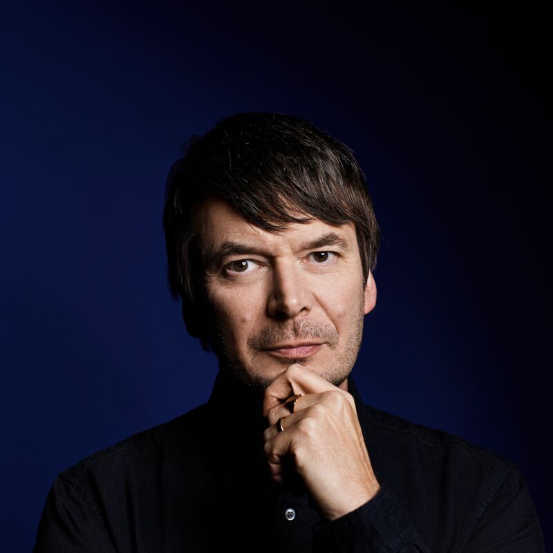 Headshot of the author Ian Rankin, he wears a black shirt and has his hand raised to his chin thoughtfully.