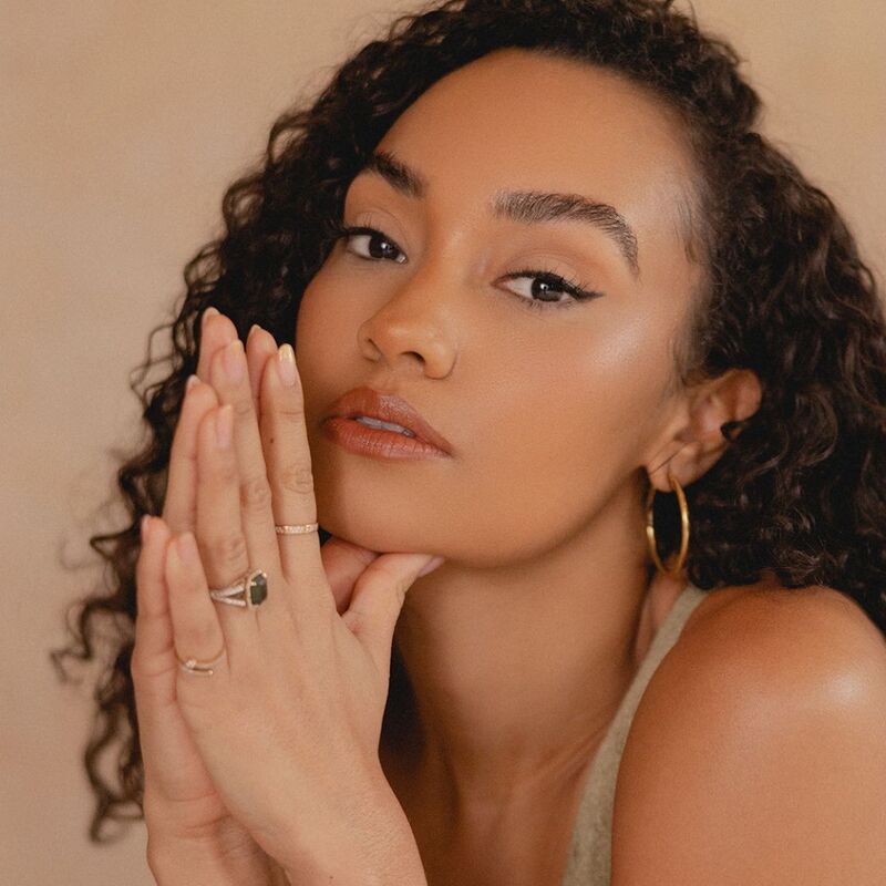 Leigh-Anne has her hands pressed together in a prayer position and resting to the side of her face. She has long, dark, curly hair and brown eyes and wears a camel coloured vest.