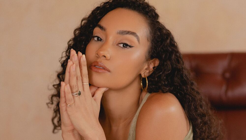 Leigh-Anne has her hands pressed together in a prayer position and resting to the side of her face. She has long curly dark hair and brown eyes and wears a camel coloured vest top.