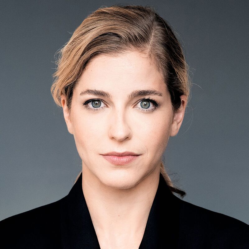 Head of shoulders shot of conductor Karina Canellakis wearing a black blazer against a grey backdrop