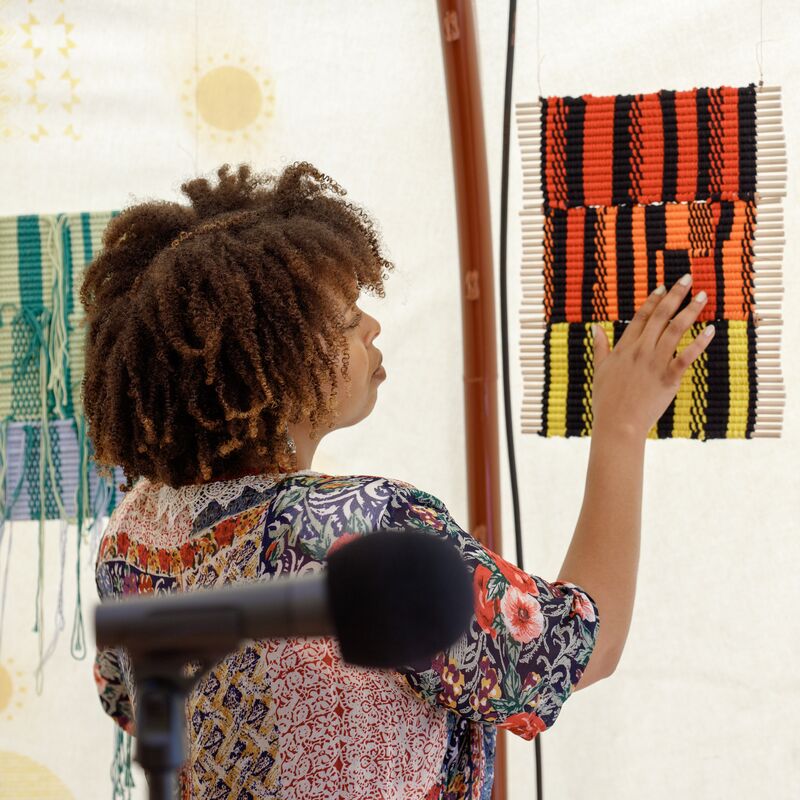 A Black woman stands in front of 3 woven segments of material and her hand reaches out to touch the one in front of her.