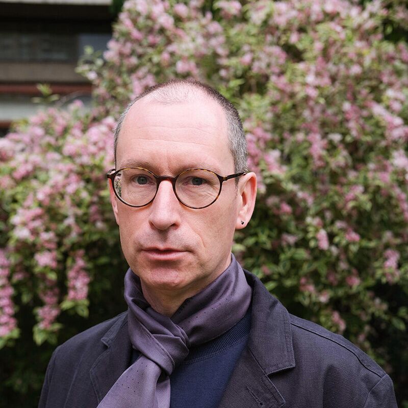 Brian Dillon faces the camera wearing a glasses and a scarf standing outside in front of a flowering bush