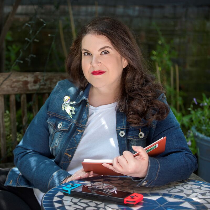 Author photo of Naomi Alderman reading on a park bench wearing a denim jacket and white t-shirt. She has long brown hair and red lipstick