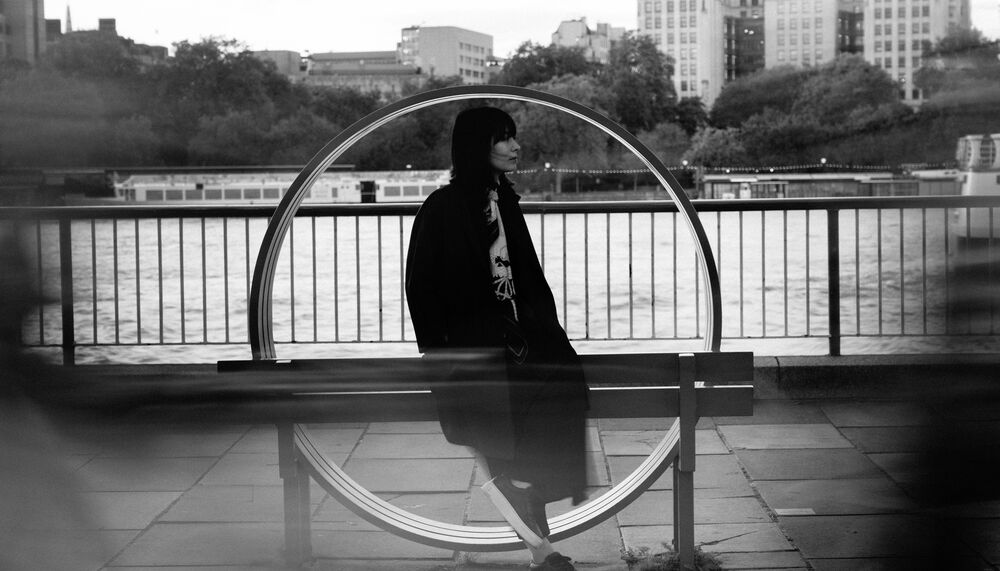 Pianist Alice Sara Ott, a young woman with short dark hair leans on a circular bench in front of the River Thames