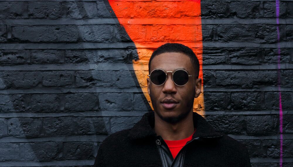 Benna is photographed against a brick wall that is painted black with a vivid orange stripe down it. Benna wears dark, round sunglasses, a black jacket and an orange tshirt underneath to match his surroundings.