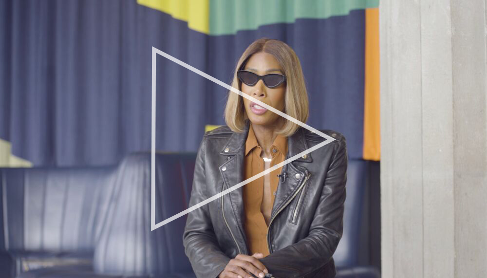Honey Dijon, a Black woman with short light brown hair, wearing sunglasses and a leather jacket speaks to camera whilst seated in the Queen Elizabeth Hall Foyer