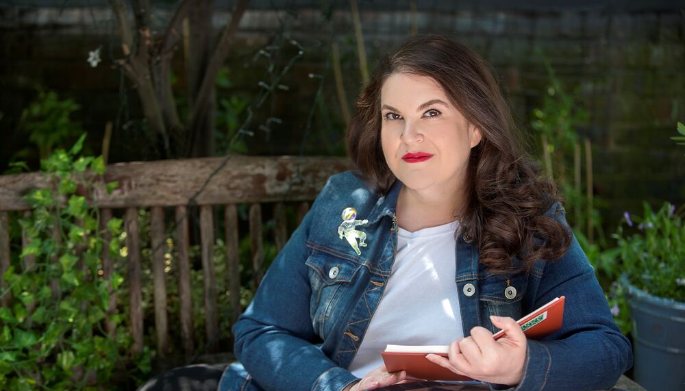 Author photo of Naomi Alderman reading on a park bench wearing a denim jacket and white tshirt. She has long brown hair and red lipstick.