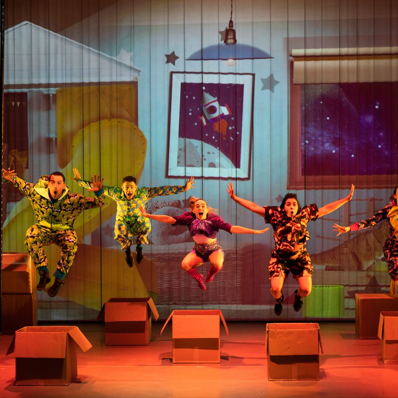 Five actors wearing pyjamas jumping over cardboard boxes.