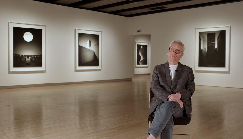 Hiroshi Sugimoto, an older Japanese man with white hair, wearing glasses and a grey suit jacket over a white t-shirt sits in the Hayward Gallery, behind him are a number of his art works on the walls of the Hayward Gallery's Hiroshi Sugimoto exhibition
