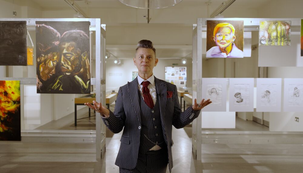 Joelle Taylor, a middle-aged woman who wears a three piece suit and her grey hair styled in a quiff stands among the artworks, encased in vitrines, of the 2023 Koestler Arts exhibition IN CASE OF EMERGENCY, which she curated