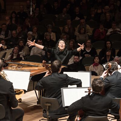 Mitsuko Uchida with open arms facing the Mahler Chamber Orchestra. Audience are sitting behind her in the shot