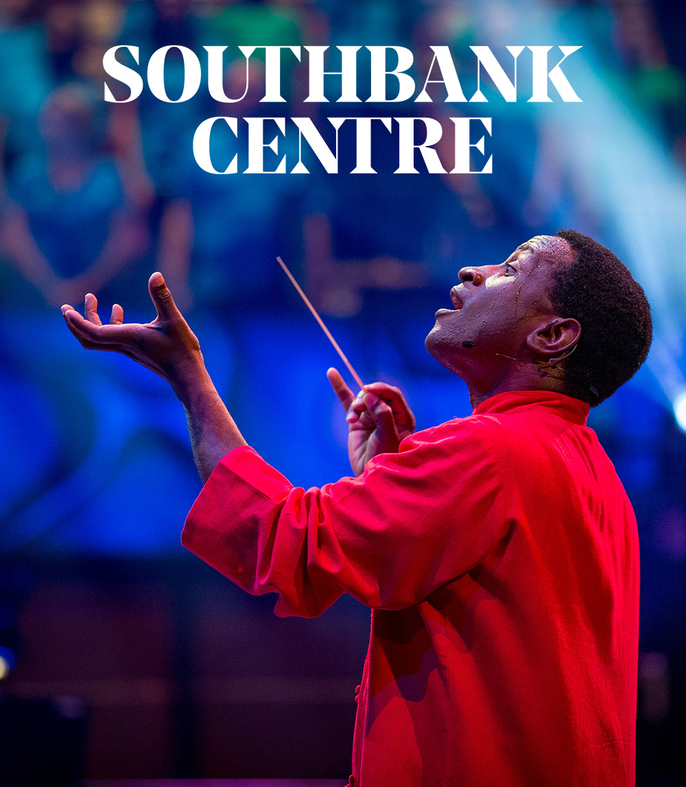 Head and shoulders shot of conductor Kwamé Ryan on stage, mid performance. He wears a red shirt, a baton in his right hand