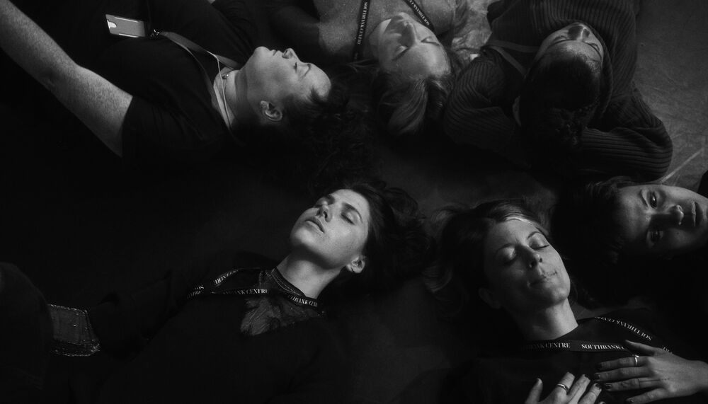 6 people lay on the floor with their eyes closed in black and white.