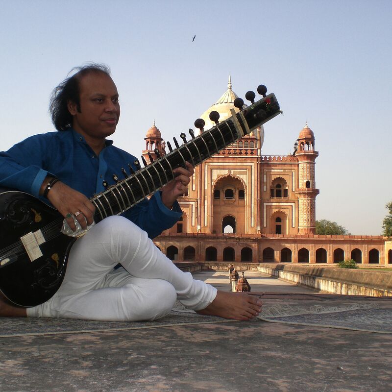 Nishat Khan sitting on the ground in front of a temple playing a sitar.