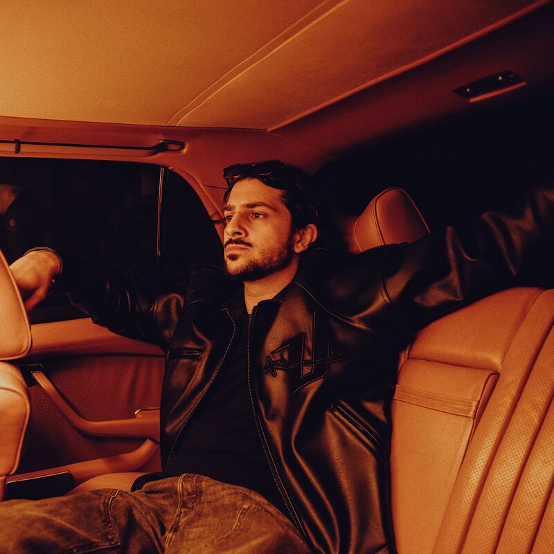 A man wearing a leather jacket sat in the back seat of a car and lit in moody red lighting