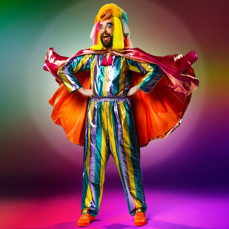 Prancer the Dancer stands with their hand on their hips with their cape flying behind them. They are wearing a purple, turquoise and yellow shiny striped jumpsuit with bright orange cape and spray painted orange high top trainers. Prancer’s hair is orange, yellow, pink and white. It is quiffed at the front with pom poms and multi coloured sections