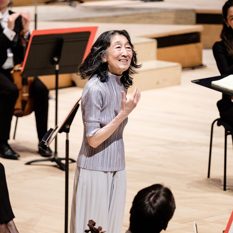 Pianist Mitsuko Uchida standing on stage with the Philharmonia at the Royal Festival Hall, smiling to acknowledge applause, wearing a silver pleated outfit