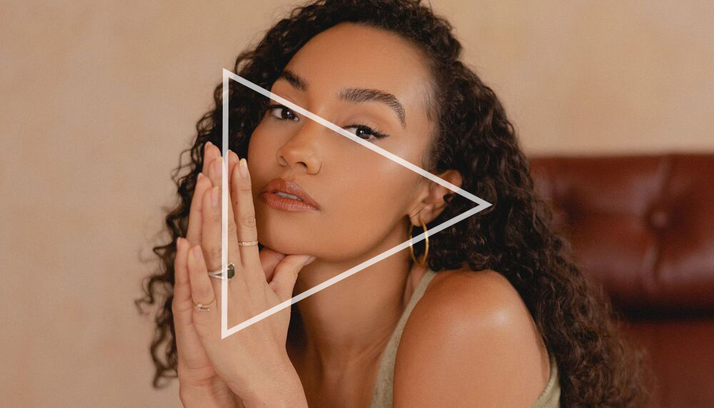 Leigh-Anne has her hands pressed together in a prayer position and resting to the side of her face. She has long curly dark hair and brown eyes and wears a camel coloured vest top.
