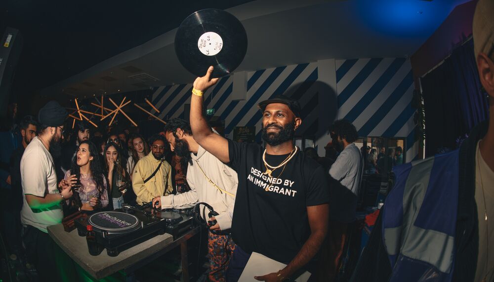 DJ stands holding a vinyl record aloft in a busy club, with dancers behind him.