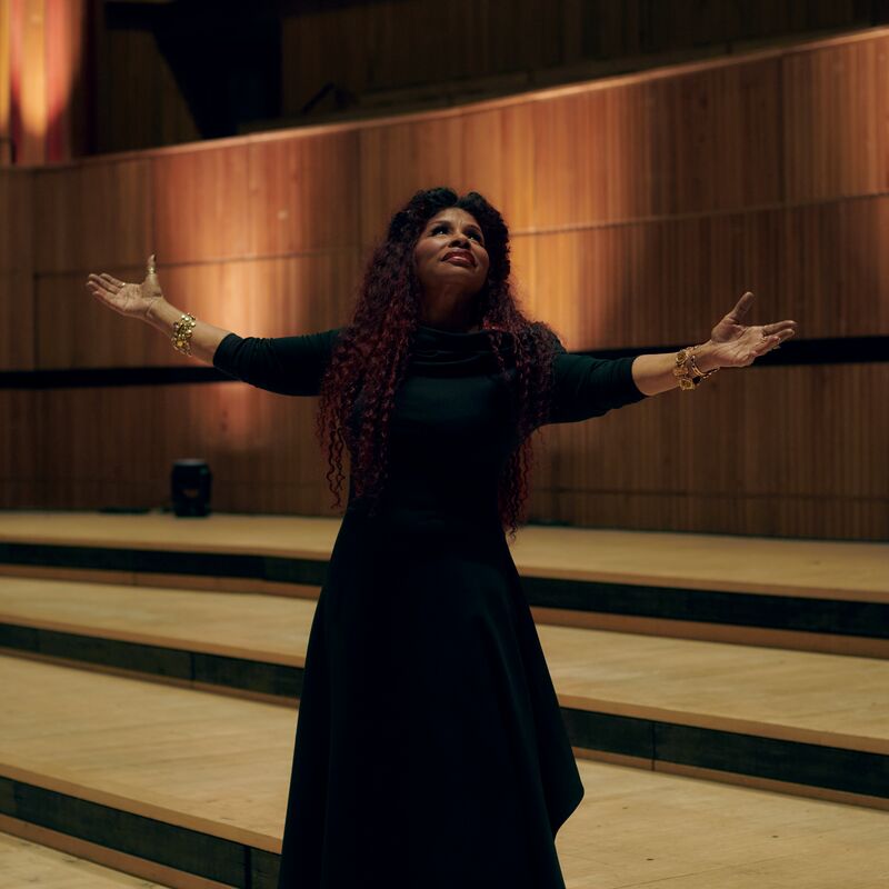 Chaka Khan standing on stage with her arms outstretched wide and wearing a long black dress.
