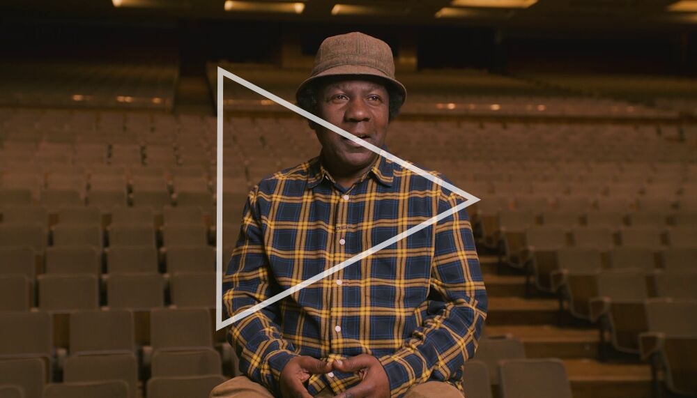 The DJ Norman Jay in the Royal Festival Hall. Jay sits on the stage with the empty seats of the auditorium behind him. Jay, an older Black man wears a plaid check shirt and a rimmed hat