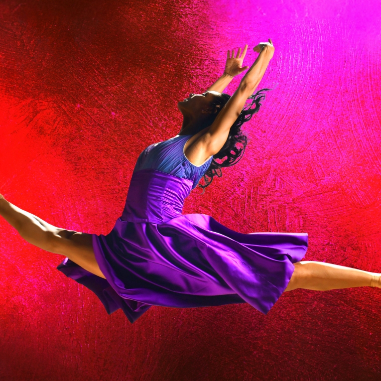 Ballet dancer wearing a purple dress is mid air in a pose against a red and pink background